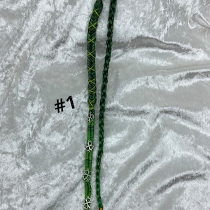 May include: Two green hair wraps, one braided and one wrapped with green thread and yellow crisscross stitching. The wrapped wrap has silver clover charms. The number "#1" is printed in black on the left side.