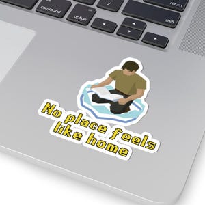 May include: A sticker featuring a pixelated illustration of a person reading on a floating ice floe. The sticker has the text "No place feels like home" in yellow. The sticker is on a laptop.