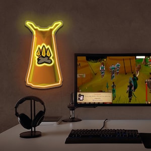 Może przedstawiać: Neonowy znak w kształcie peleryny z symbolem ogniska. Znak jest żółty i pomarańczowy, zamontowany na ciemnej ścianie nad biurkiem. Monitor komputera wyświetla scenę z gry wideo, ze słuchawkami i klawiaturą na biurku.