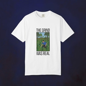 May include: White t-shirt with a graphic that reads "THE GRIND WAS REAL." The graphic depicts a person working in a field. The shirt is made of a soft material and has a crew neck.