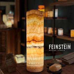 Puede incluir: Lámpara cilíndrica de piedra en capas, con bandas de color naranja, amarillo y marrón. La lámpara está iluminada desde dentro. El texto "FEINSTEIN design studio berlin" es visible. La lámpara mide unos 30 cm de altura.