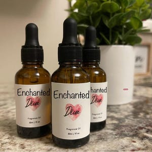 May include: Three amber glass bottles of Enchanted Diva fragrance oil with black droppers. Each bottle has a white label with the words "Enchanted Diva" and a watercolor heart. The bottles are 1 fl oz / 30 ml.