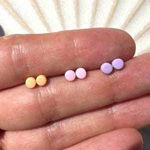 May include: Three pairs of small, round stud earrings in pastel shades: peach, pink, and lavender. The earrings are shown on a finger, highlighting their size and colour variations. These minimalist earrings are suitable for everyday wear.