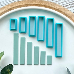 May include: A selection of teal and light green rectangular cookie cutters displayed on a white and wooden tray. The cutters vary in size, with solid and frame-style designs. The arrangement suggests a baking or crafting theme.