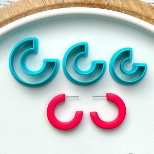 May include: A collection of turquoise letter-shaped polymer clay cutters and a pair of bright pink hoop earrings. The cutters are in three sizes, arranged on a white plate, with a wooden tray in the background.
