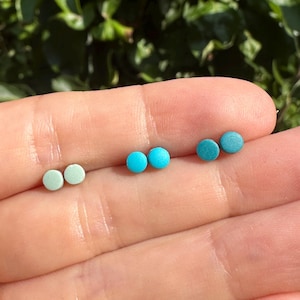 May include: Three pairs of small, round stud earrings in shades of blue and green. The earrings are displayed in a hand against a blurred green backdrop, highlighting the different colours and styles of the jewellery.