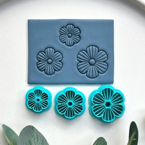 May include: A collection of turquoise flower-shaped cookie cutters and a blue rectangular mold with flower imprints. The cutters and mold are displayed on a white surface, with green foliage. The cutters are for baking, and the mold is for creating flower shapes.