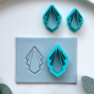 May include: A set of light blue, geometric-shaped cookie cutters. The set includes two individual cutters and a matching impression stamp. The cutters and stamp are arranged on a light blue surface, with a white background.
