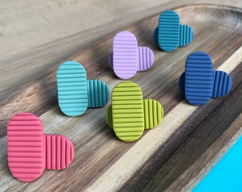 May include: Six pairs of colorful, striped, oval-shaped earrings. The earrings are arranged in a row on a wooden surface.