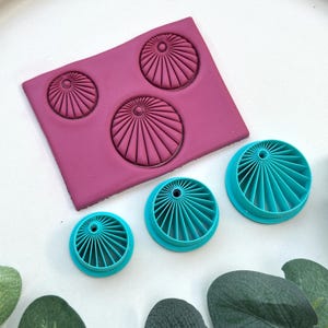 May include: A close-up of polymer clay tools. A rectangular magenta clay block has three circular impressions. Three teal-coloured circular cutters are also visible. The tools have a radial design.