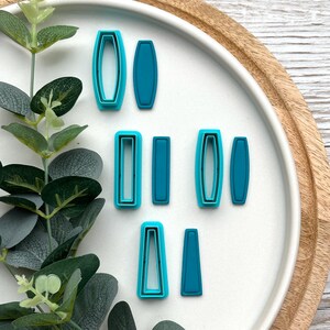 Könnte beinhalten: Eine Sammlung türkisfarbener Polymer Clay Ausstecher in verschiedenen rechteckigen und ovalen Formen. Die Ausstecher sind auf einem weißen Teller mit Holzrand angeordnet, neben einer grünen Pflanze mit Blättern.