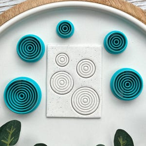 Könnte beinhalten: Eine Sammlung türkisfarbener, kreisförmiger Ausstecher mit konzentrischen Ringmustern. Die Ausstecher sind um ein Quadrat aus Teig mit passenden geprägten Kreisen angeordnet. Die Artikel werden auf einem weißen Teller mit Holzrand präsentiert.