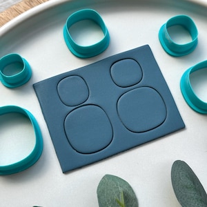 May include: A teal cookie cutter set with a dark blue rectangular piece featuring four rounded square cutouts. Several teal ring-shaped cutters surround the rectangular piece. The items are arranged on a white plate, with green leaves in the background.