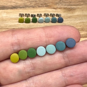 May include: A selection of seven circular stud earrings in a gradient of colours, ranging from yellow-green to dark blue. The earrings are shown on a hand, with the posts and backs visible. The background is a light wood surface.