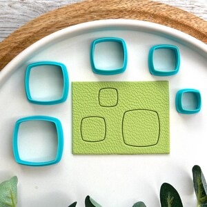 May include: A selection of turquoise square cookie cutters with rounded corners, alongside a textured green rectangle featuring square cutouts. The items are presented on a white plate with a wooden rim.