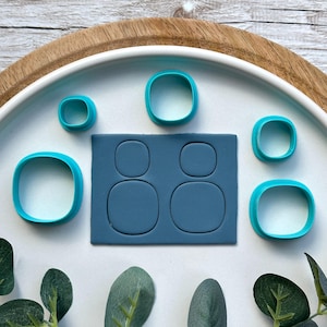 May include: A collection of teal-colored, rounded square cookie cutters and a matching clay mold. The cutters vary in size, with a rectangular mold featuring four rounded square impressions. The items are arranged on a white surface.