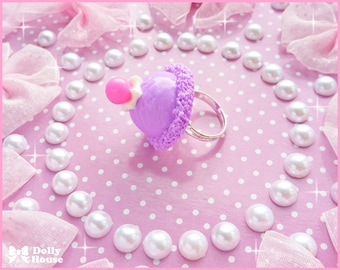 May include: A purple and pink ring with a cupcake design. The ring is surrounded by white pearls on a pink background with white polka dots.