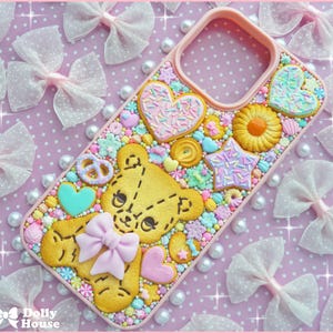 May include: A pink phone case decorated with a yellow teddy bear, hearts, stars, and various cookie-shaped embellishments. The case is adorned with pastel-colored sprinkles and a pink bow. The background features white polka-dot bows and pearls.