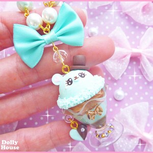 May include: A mint green and brown charm necklace with a cute ice cream cone charm featuring a bear wearing a top hat. The charm is surrounded by a mint green bow and a pearl and gold chain.