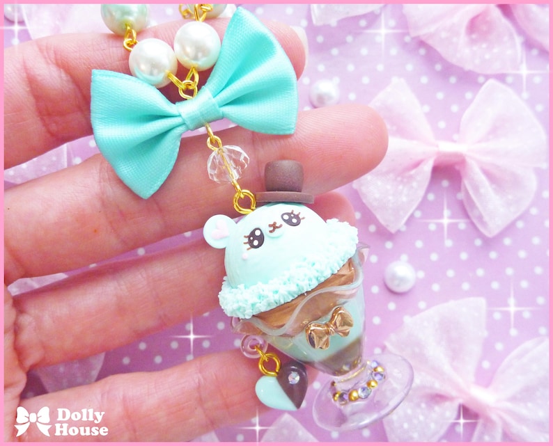 May include: A mint green and brown charm necklace with a cute ice cream cone charm featuring a bear wearing a top hat. The charm is surrounded by a mint green bow and a pearl and gold chain.