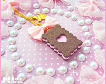 May include: A pink and brown chocolate-covered ice cream sandwich charm necklace with a gold chain and a pink bow. The charm is on a pink background with white polka dots.