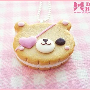 Puede incluir: Una galleta con forma de oso rosa y marr&oacute;n con un coraz&oacute;n rosa y un parche negro para el ojo. La galleta est&aacute; sobre un fondo blanco y rosa y est&aacute; unida a una cadena plateada.