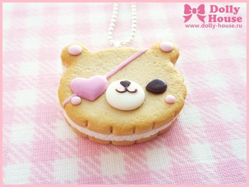 Puede incluir: Una galleta con forma de oso rosa y marr&oacute;n con un coraz&oacute;n rosa y un parche negro para el ojo. La galleta est&aacute; sobre un fondo blanco y rosa y est&aacute; unida a una cadena plateada.