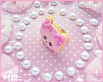 May include: A pink and white bear-shaped cookie with a pink bow on its head. The cookie is surrounded by white pearls on a pink background.