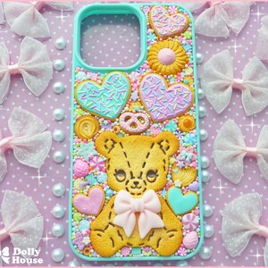 Op de afbeelding: Een turquoise telefoonhoesje versierd met een gele teddybeer, hartvormige koekjes en snoepjes. De hoes heeft een lichtroze strik en is omgeven door roze strikken en parels.