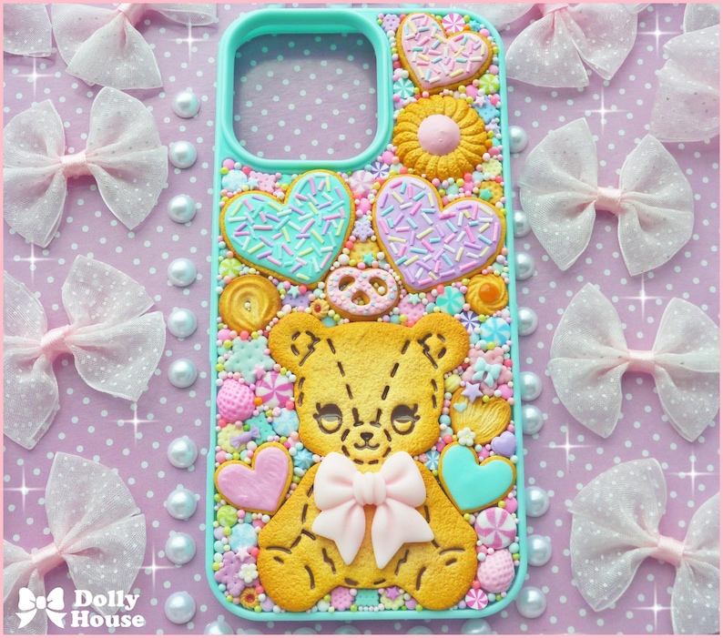 Op de afbeelding: Een turquoise telefoonhoesje versierd met een gele teddybeer, hartvormige koekjes en snoepjes. De hoes heeft een lichtroze strik en is omgeven door roze strikken en parels.