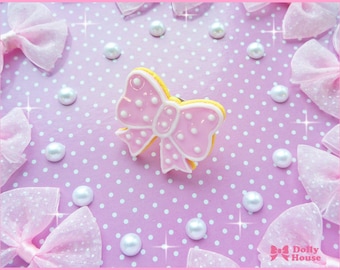 May include: A pink bow-shaped cookie with white polka dots on a pink background with white polka dots. The cookie is on a white surface.