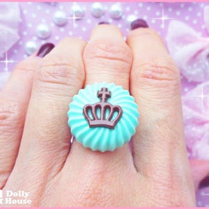 K&ouml;nnte beinhalten: Ein t&uuml;rkisblauer Ring mit einem braunen Kronendesign. Der Ring befindet sich an einem Finger und der Hintergrund ist ein rosa-wei&szlig;es Punkte-Muster.