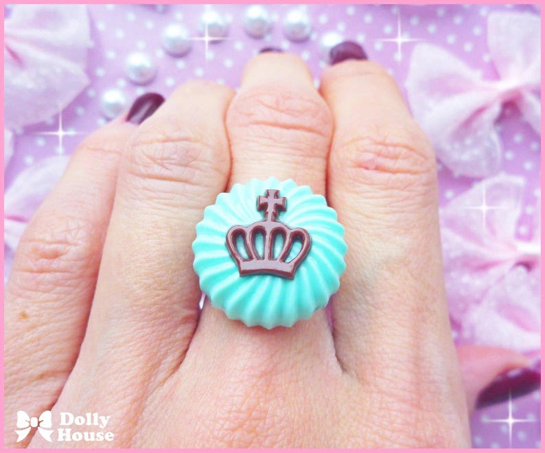 K&ouml;nnte beinhalten: Ein t&uuml;rkisblauer Ring mit einem braunen Kronendesign. Der Ring befindet sich an einem Finger und der Hintergrund ist ein rosa-wei&szlig;es Punkte-Muster.
