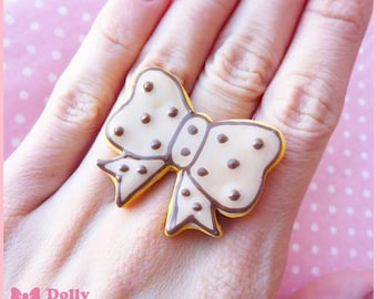 May include: A white bow-shaped ring with brown polka dots. The ring is made of polymer clay and is a unique and whimsical accessory.