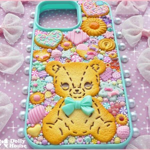 Op de afbeelding: Een turquoise telefoonhoesje versierd met een verscheidenheid aan kleurrijke snoepjes en koekjes. Het ontwerp omvat een grote gele teddybeerkoek met een turquoise strik, omringd door harten, sterren en andere zoete lekkernijen. De achtergrond is een roze stippenpatroon.