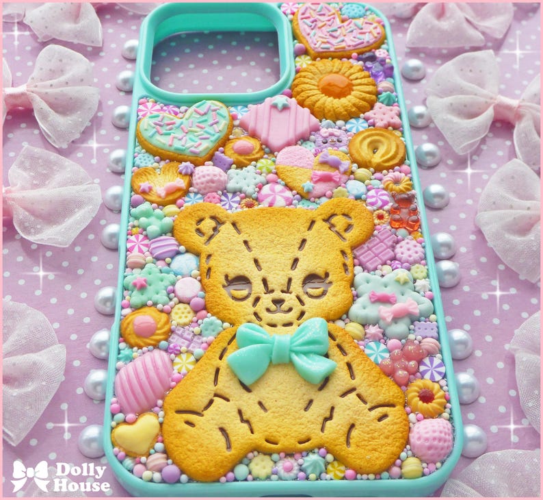 Op de afbeelding: Een turquoise telefoonhoesje versierd met een verscheidenheid aan kleurrijke snoepjes en koekjes. Het ontwerp omvat een grote gele teddybeerkoek met een turquoise strik, omringd door harten, sterren en andere zoete lekkernijen. De achtergrond is een roze stippenpatroon.