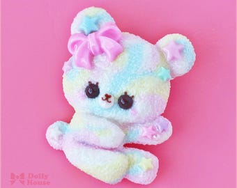 May include: A pastel rainbow colored bear charm with pink, blue, and yellow swirls. The bear has a pink bow with a blue star on top and a blue star on its ear. The bear has brown eyes and a white nose.