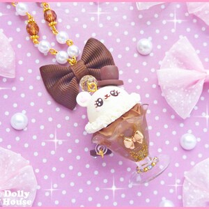 May include: A brown and white chocolate ice cream sundae charm necklace with a cute bunny face, a brown bow, and a gold top hat. The necklace is on a pink background with white polka dots.