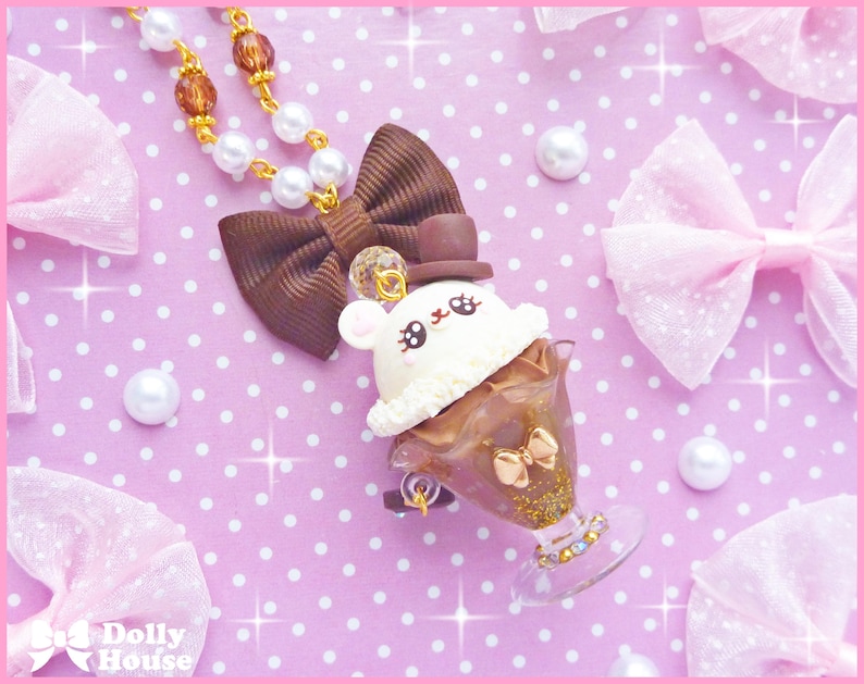 May include: A brown and white chocolate ice cream sundae charm necklace with a cute bunny face, a brown bow, and a gold top hat. The necklace is on a pink background with white polka dots.
