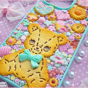 Op de afbeelding: Een decoratieve telefoonhoes met een teddybeerontwerp gemaakt van koekjes en snoep. De hoes is versierd met diverse snoepjes in pastelkleuren, waaronder hartjes, sterren en strikjes. De hoes heeft een lichtblauwe rand en staat tegen een roze stippenachtergrond.