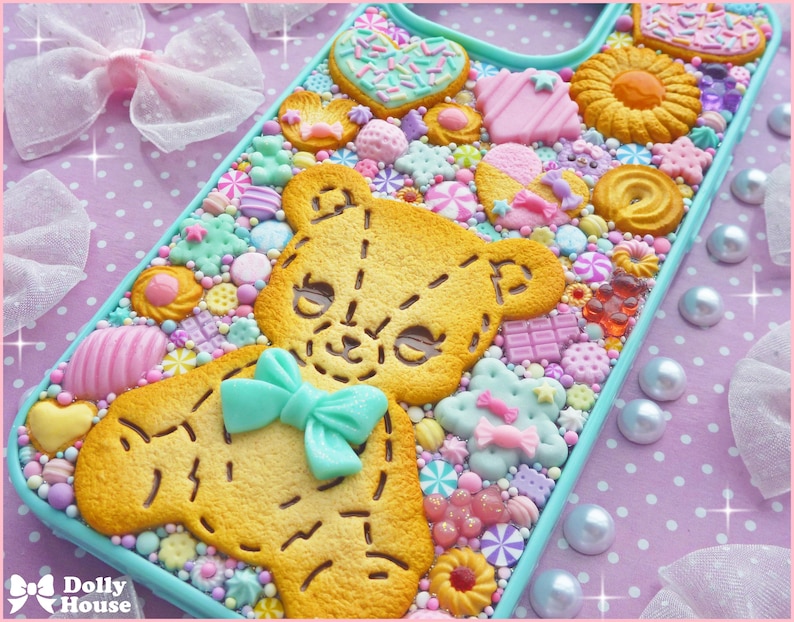 Op de afbeelding: Een decoratieve telefoonhoes met een teddybeerontwerp gemaakt van koekjes en snoep. De hoes is versierd met diverse snoepjes in pastelkleuren, waaronder hartjes, sterren en strikjes. De hoes heeft een lichtblauwe rand en staat tegen een roze stippenachtergrond.