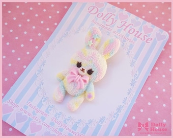 May include: A pastel rainbow colored bunny brooch with a pink bow and a sparkly star on its head. The brooch is handmade and is displayed on a pink and blue striped background with white polka dots. The text "Dolly House handmade accessories" is visible in the top left corner of the image.