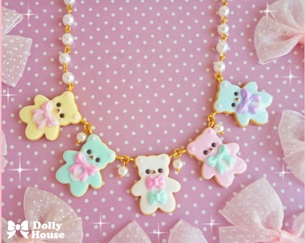 Op de afbeelding: Een pastelkleurige ketting met een gouden ketting en zes berenbedels. De beren zijn versierd met pastelkleurige glazuur en strikjes. De ketting ligt op een roze achtergrond met witte stippen.