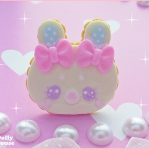 Può includere: Un biscotto a forma di coniglio bianco e rosa con un fiocco rosa e pois blu. Il biscotto è su uno sfondo rosa con perle bianche.