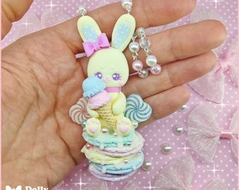 May include: A pastel yellow clay bunny pendant featuring a pink bow, holding a blue and pink ice cream cone. The bunny sits atop a stack of pastel-colored macarons, accented with white pearls and a silver chain with beads.