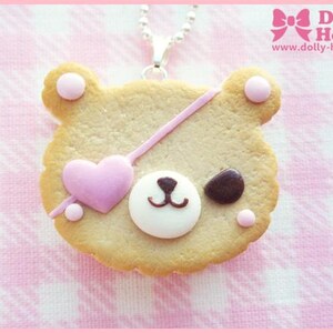 Puede incluir: Una galleta con forma de oso marr&oacute;n con glaseado rosa y blanco, un coraz&oacute;n rosa y un parche en el ojo marr&oacute;n. La galleta est&aacute; sobre un fondo a cuadros rosa y blanco y est&aacute; unida a una cadena plateada.