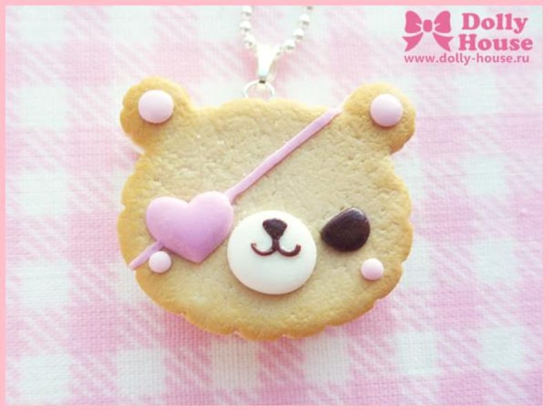 Puede incluir: Una galleta con forma de oso marr&oacute;n con glaseado rosa y blanco, un coraz&oacute;n rosa y un parche en el ojo marr&oacute;n. La galleta est&aacute; sobre un fondo a cuadros rosa y blanco y est&aacute; unida a una cadena plateada.