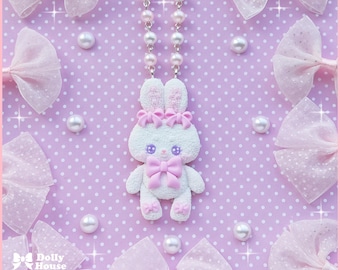 May include: A white bunny pendant necklace with pink bows and pearl accents. The bunny has purple eyes and is on a pearl chain. The background is a light purple dotted surface with pink bows and pearls.