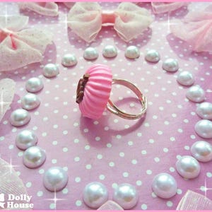 K&ouml;nnte beinhalten: Ein rosafarbener Cupcake-Ring mit einem silbernen Band. Der Ring ist von wei&szlig;en Perlen umgeben auf einem rosa Hintergrund mit wei&szlig;en Punkten.