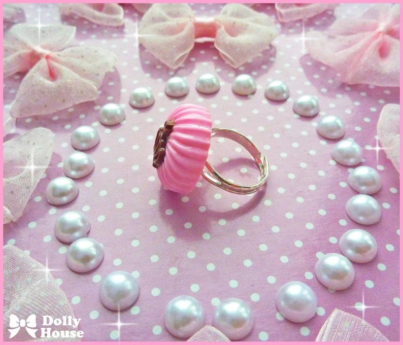 K&ouml;nnte beinhalten: Ein rosafarbener Cupcake-Ring mit einem silbernen Band. Der Ring ist von wei&szlig;en Perlen umgeben auf einem rosa Hintergrund mit wei&szlig;en Punkten.